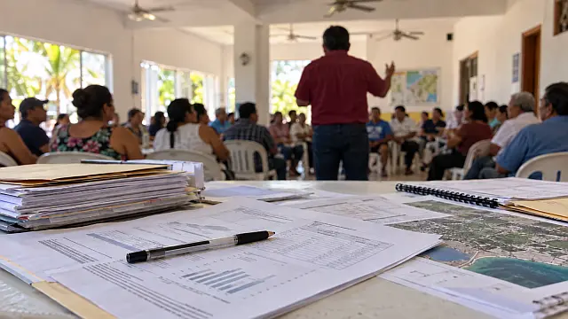 Documentos administrativos en primer plano durante una reunión comunitaria, imagen ilustrativa de la rendición de cuentas y las aspiraciones políticas.