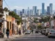 Calle de una zona popular de México, con viviendas deterioradas y pequeños comercios en primer plano y modernos rascacielos al fondo, reflejo de la desigualdad económica.